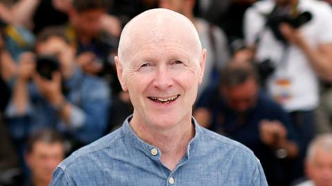 Paul Laverty smiling straight at the camera. He is wearing a light blue shirt which has the top button undone. He is bald. In the background behind him is a large group of photographers who are blurred.
