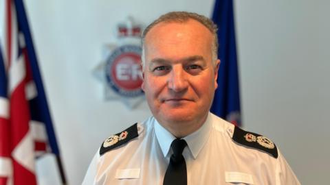 Greater Manchester Police's Chief Constable Stephen Watson, who has short, grey hair, is photographed wearing his uniform. He is standing in front of his force's logo and two flags.