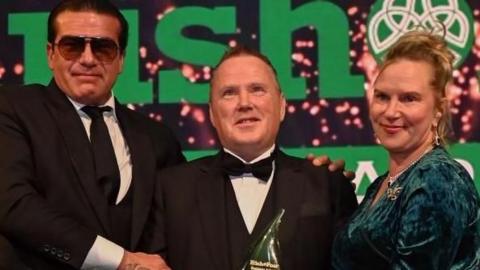 Billy McAllister stands in the middle of three people as he is presented with the 2023 Inspiration in the Community Award at the Irish Post Awards.