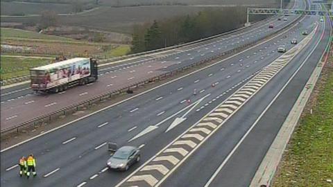 The M11, with two people walking on it with a high-viz jackets on, there are a few cars in one lane and a  lorry in the other. I shows traffic cones, and several lanes of road. 