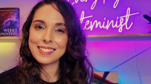 Chloe Lipton is looking and smiling directly at the camera. She is wearing a black T-shirt and has long, brown hair. She is sitting in front of a shelving unit which is lit with brightly coloured lights and a neon sign which reads "Everyday Steminist" in pink, illuminated letters.