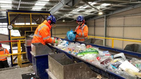 Two men working inside a recycling centre. The men are either side of a conveyor belt carrying plastic waste such as carrier bags, milk cartons and bottles. The workers are wearing orange high visability jackets, blue hardhats and red ear defenders.