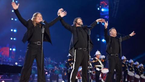 Mark Owen, Howard Donald and Gary Barlow of the band take that perform on a stage. They each stand in front of microphones on stands and have their arms outstretched. 
