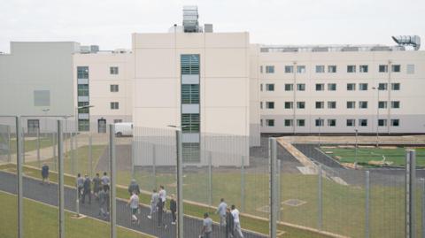 The exterior of HMP Five Wells, a white panelled building, with several inmates walking around the gated perimeter. There is a high metal fence.