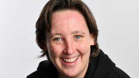 Mhairi Black, a young woman with short loose brown hair, smiles into the camera against a grey backdrop. She is wearing a black hoody.