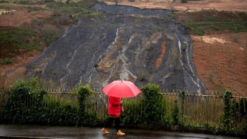 Woman with umbrella walks past coal tip landslide in Tylorstown, Rhondda Cynon Taf