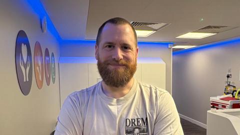 Paul Sewry smiling at the camera and stood inside the new centre. He has a large beard and is wearing a grey top. On the white wall to his left there are circle signs with different logos inside.