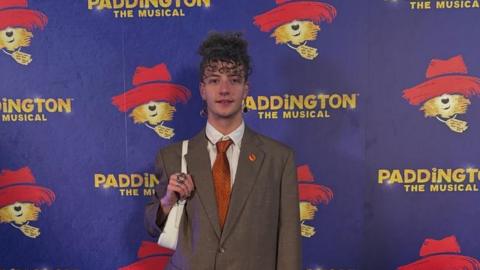 Rhys is a young man with curly brown hair. He is wearing a brown suit with orange tie and white shirt and has a pendant earing in each ear. He is clutching the white strap of a bag over his shoulder and is stood infront of a wall which has been covered in a large 'Paddington the muscial' poster.