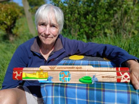 Lesley Sleight, wearing a blue jumper, leans on a table where the baton rests. It is a three-sided wooden baton, featuring coloured paintings of Manx heritage sites, and cultural events. 
