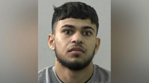 Mugshot of a man with a black beard and hair and wearing a grey sweatshirt