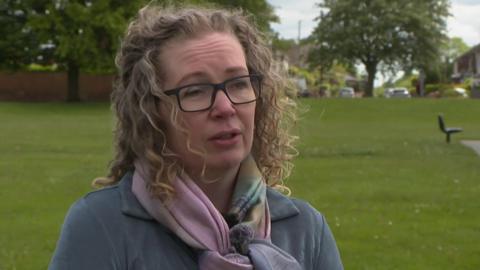 Andrea Jack has long blonde curly hair and is wearing dark-framed glasses, a blue jumper and a pink scarf. She is standing in a park with trees in the distance.