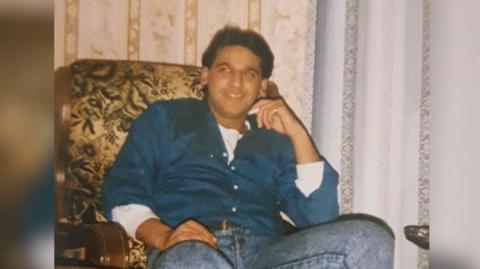 A photograph of Ethsham Ul-Haq Ghafoor wearing blue denim jeans and a blue shirt sitting on an armchair  smiling. 