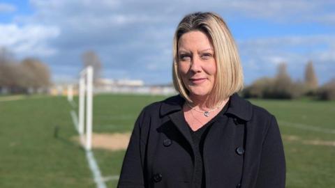 Natalie Webb has blonde hair cut into a bob. She is wearing a black wool coat over a black sweater.  Behind her are white football posts, without nets, on a worn playing field.