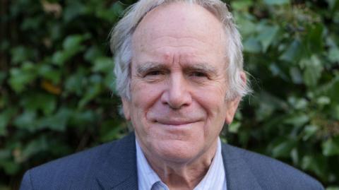 Professional picture of Adrian Birch, photographed wearing a grey suit jacket and light coloured shirt against a leafy green background. 