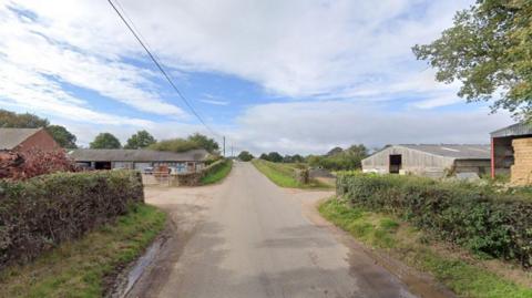 A road with agricultural buildings either side. There is a small grass verge are hedgerows lining the road and a telephone wire running above the left hand side.