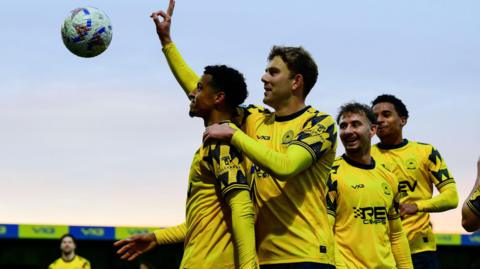 Louis Dennis (left) celebrates his hat-trick for Torquay United against Eastbourne, surrounded by teammates.
