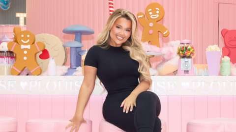 A promotional shot of a smiling Paige Louise Williams. She has long blonde hair and wears black trousers and t-shirt. She is pictured sitting on a pink bar stool in front of an all pink bar decorated with P.Louise branding.