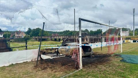 An image of a white car that has been completely burnt out, it is sitting inside a goal post on a football pitch. The goal post and its net are badly damaged, and a patch of grass around the vehicle scorched. The hoarding around the pitch is also damaged where the car has driven into it. 