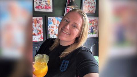 A women smiling and looking directly at the camera. She is wearing a black England football shirt and holding a glass with an orange drink in it. 