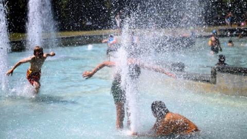 Teenage boys playing in a large fountain of a splash park. Their faces are obscured from the camera. One boy has his face in a large water fountain spray. It is a bright sunny day.