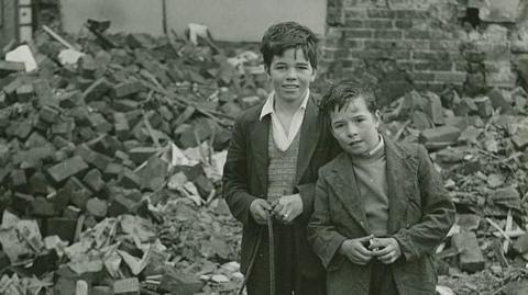 An image of two children from the 1960s stood in front of a pile of bricks next to a damaged wall.