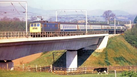 An old colour photograph of a blue and yellow train passing over the old Clifton Bridge. The grey concrete structure rises over a grassy valley. A cow is grazing on the nearside, behind a wooden fence.