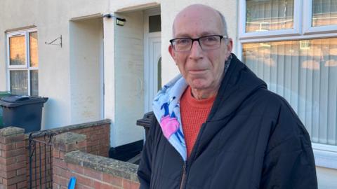 An older man with short grey hair at the back of his scalp, black rectangular rimmed glasses, a red jumper and a black hoodie with a fleecy, patterned interior stands outside of his terraced home in a city suburb on a sunny winter morning,