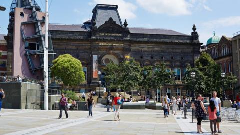 The front of Leeds City Museum. It is a sunny day, with people pictured walking in front of the building.