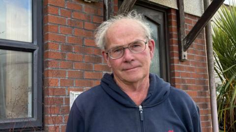 Veryan parish councillor Luke Dunstone stood outside the house. He is wearing glasses and has a zip-up hoodie on. 