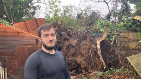 Curtis Carvell, wearing a black top, stood in front of the roots of a large tree which has fallen down