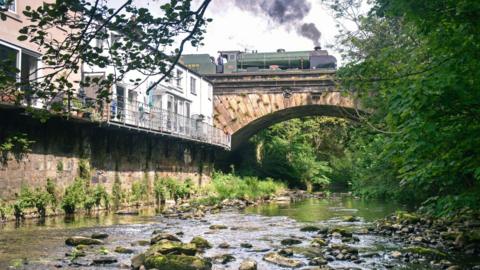 A picture of an old bridge with a green steam train going over it with a river below 