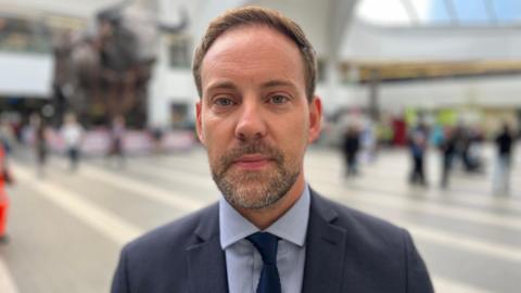 A man with short brown hair is standing in a train station. He has grey and brown facial hair and is wearing a navy suit jacket, blue shirt and navy blue tie.