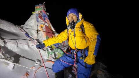 A man in bright yellow and blue climbing gear stands in front of an icy peak that is covered in prayer flags