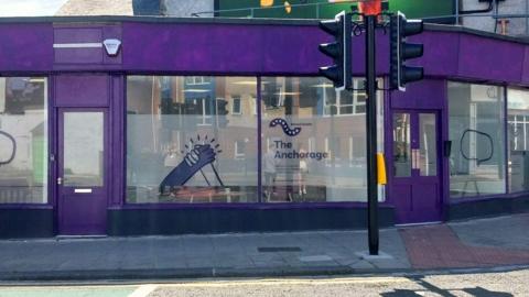 The Anchorage. The drop-in centre is a wide former retail building. It doors and panelling has been painted purple and it has large glass windows all the way around. The middle section of each window has been frosted and decorated with signage for the centre including two hands holding each other.