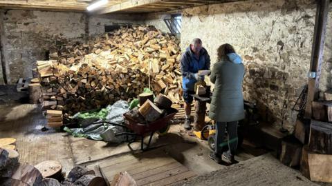 A big pile of logs and two people working together to cut them, with a loaded wheelbarrow to one side 