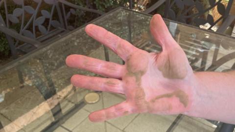 The hand of Sandra Caller has dust on it after she swipes her garden table with it. Her hand is raised to the camera, with a glass garden table in the background