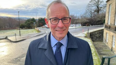 Prof Alan McNeill stands on the pavement with an old office building to his left and a grassy hill to his right and behind them. He is wearing a navy coat and a blue shirt and tie and glasses.