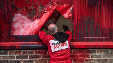 A member of the Palestine Action protest group dressed in red overalls, throwing red paint over the Elbit building and smashing windows