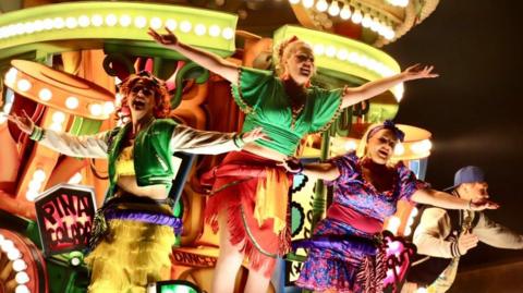 A lit-up carnival cart with three women and a man standing on it in colourful costumes. They are holding up their arms and smiling.