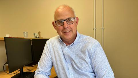 A man sits next to two black computer screens. He has short grey hair and wears black glasses. He wears a light blue patterned shirt and jeans.