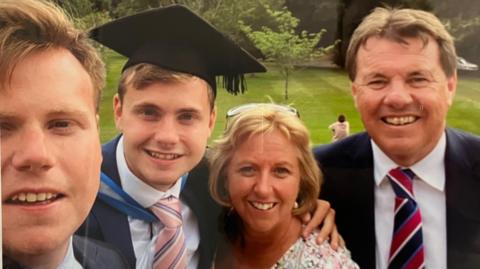 Jack O'Sullivan, pictured on his graduation day with his family.  His brother is on the left of the frame. He has fair short hair and is smiling. Jack is next to him. He also has short fair hair and is wearing a graduation cap and gown over a white shirt and pink and blue striped tie. Catherine is next to him. She has short blonde hair and has glasses on her head. She is looking at the camera and smiling. Alan O'Sullivan is on the right and is wearing a suit.