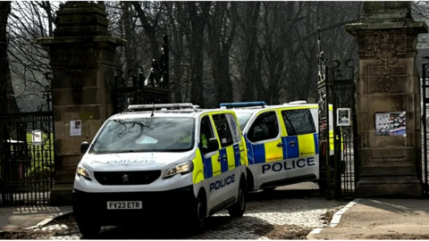 Two police vans parked between two large black iron gates and a wall- the entrance to a park