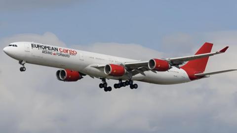 A white and red European Cargo plane in flight. Its wheels are down and there are clouds in the background. The body of the aircraft - which as a converted passenger plane still has windows along the side - is white. The tail is a completely red and, underneath the company name is the legend "moving forward". The aircraft has four jet engines, two on each wing, all painted the same red colour.