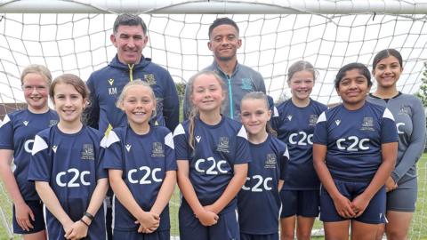 A team of young girls all lined up in front of a football goal and smiling. They are standing in front of Southend United boss Kevin Maher and captain Nathan Ralph. They are wearing blue kits.