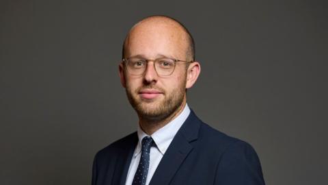 Lloyd Hatton MP - A man with a bald head and glasses looking at the camera. He has a stubbly beard. He's wearing a blue suit, with a white shirt and a blue and white dotted tie. 