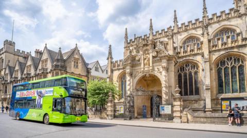 A green and blue bus is on a road with an Oxford University building in the background.