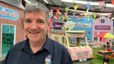 A man smiles as his stands in an indoor soft play centre. He has short grey hair and wears a black shirt with a clown badge pinned to it. Behind him are play houses, a child-sized ice cream van and rainbow-coloured tables. Bunting hangs from the ceiling.