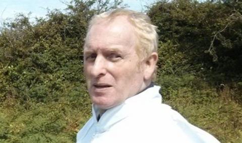 A man with blond hair, in a white jacket, standing in front of bushes