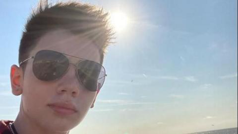 A close up image of Stefan Kluibenschadl aged 15 wearing sunglasses and looking to camera with a sunset over sea behind him  