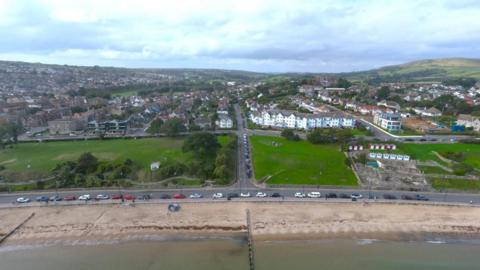 Aerial view of sea and sandy beach with cars parked along a seafront road and a large green area stretching behind.